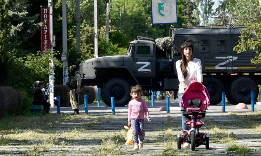A woman and child walk in a park as Russian servicemen patrol the street in Skadovsk, Kherson.
