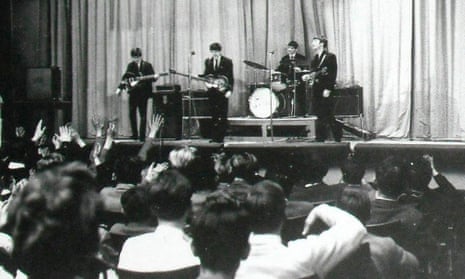Black and white photo from the back of a school auditorium showing the Beatles on stage with a young audience listening, some with their arms raised