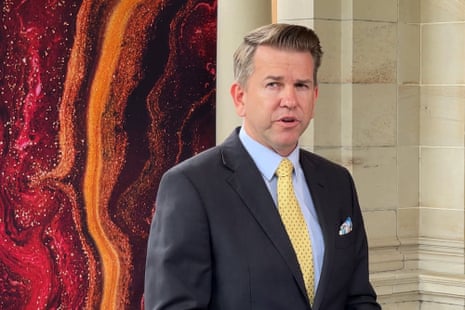 A man in a dark suit and yellow tie stands before a red background