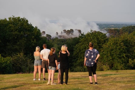Six people stand on a hill watching clouds of smoke from a fire in woodland below