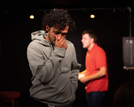 A man in a grey hoodie winces with fingers pinching his nose, with another man behind him on stage