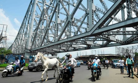 A white horse gallops alongside people on motorbikes under the steel cantilever canopy of the Howrah Bridge.