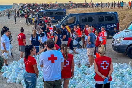 Workers in red cross clothing distributing food at a packed beach