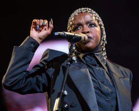 Lauryn Hill singing on stage in New York, wearing a headdress and a black jacket, holding her hand up
