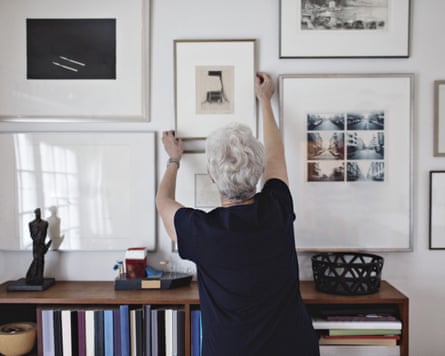 Rear view of retired senior woman adjusting picture frame on wall over bookshelf at homeAn older woman hanging pictures on a white wall