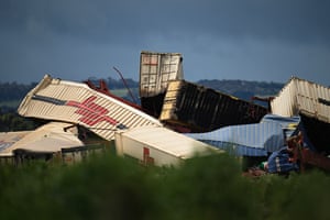 A pile of bent and damaged shipping containers lies in a field, with blurred grass in the foreground