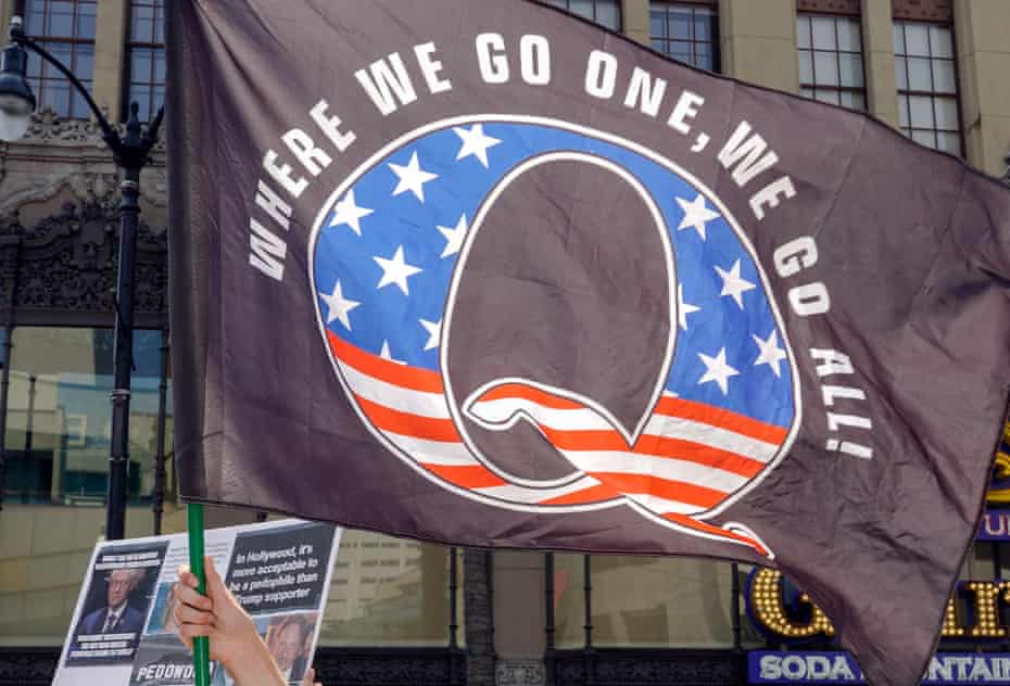 A hand holds a large flag with a stars-and-stripes adorned 'Q' on a black background. Above the 'Q' are the words 'Where we go one, we go all!'