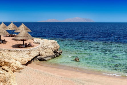 An iddylic beach scene, showing sand and azure waters, with parasols to provide shade