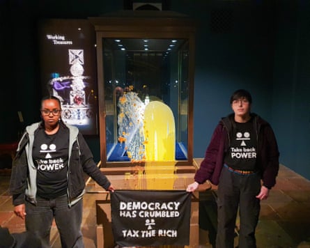 The encased crown jewels flanked by two activists holding a banner that reads ‘Democracy has crumbled. Tax the rich’.