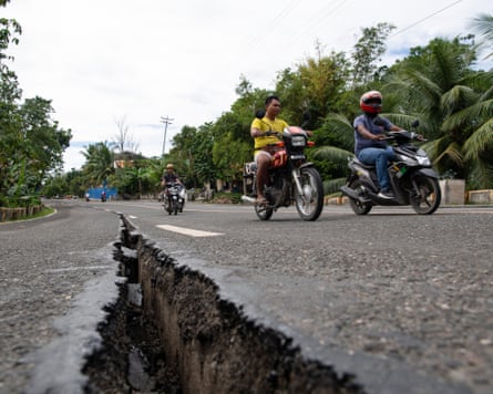 Motorcyclists pass by a large crack in the road.