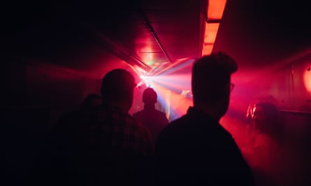 Silhouettes of people amid darkness pierced by red lights in the train’s rave carriage  