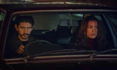 A man drives a woman, whose face is covered in blood, in a car, both of their faces with serious expressions