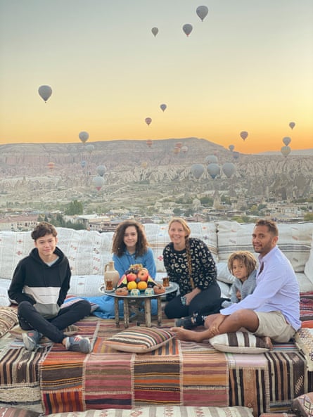 A man and woman and three children sitting around an outdoor table with fruit and drinks on it, in Turkey, with lots of hot air balloons in the background