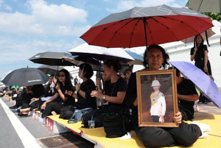 People shield themselves from the sun with umbrellas as they sit on a pavement, one holding a framed portrait of the princess