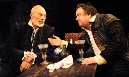Patrick Stewart, in period costume, sits at a wooden table with wine glasses on it, while Richard McCabe leans across with one arm extended.