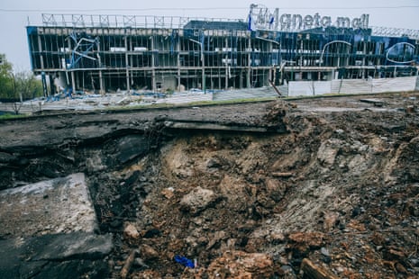 The site of a recent Russian bombing at an unused shopping mall which killed seven people. It shows a large crater in the ground and a destroyed shopping mall in the background.