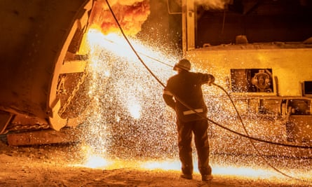 A steelworker, back to camera, is bathed in an orange glow while sparks fly as he cleans part of a blast furnace with a long lance