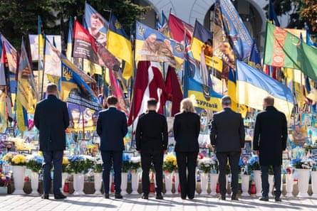 Leaders stand with backs to camera with flowers and flags in front of them on memorial