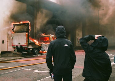 Two young people in hooded tops, seen from behind, watch a truck burn
