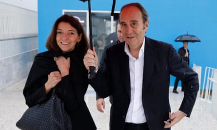 A middle-aged man and woman, both dressed in dark-coloured clothing walk along a pavement together sharing an umbrella