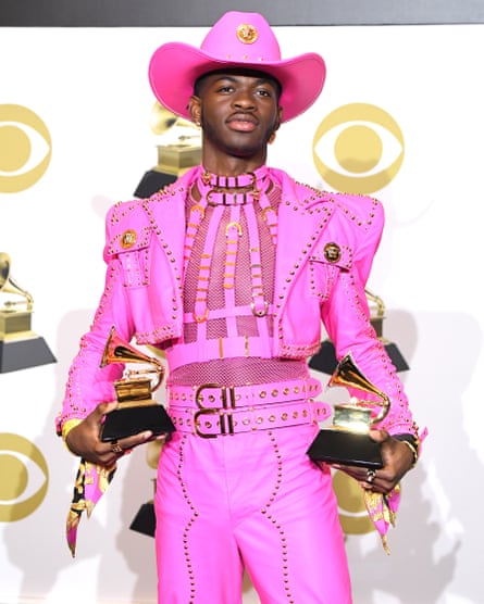 Lil Nas X wearing a leather, Barbie-pink Versace suit, with a harness, see-through mesh T-shirt and cowboy hat, holding two awards at the Grammys in January 2020