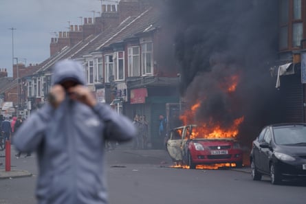 A car burns on a street in Middlesbrough with a young man covering his face with a hooded jacket