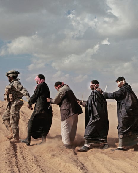 A US soldier leads four blindfolded and handcuffed men in singe file across sandy ground