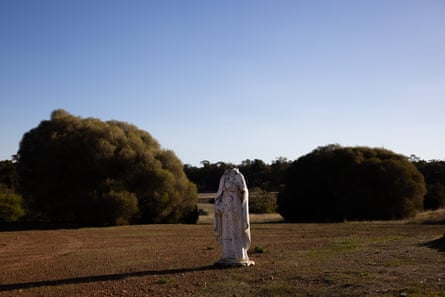 A headless statue stands in the centre of some grass with trees behind