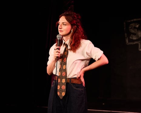 Ania Magliano holds a microphone on stage wearing a white shirt and patterned tie