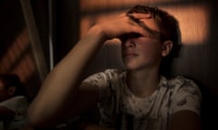 A teenage boy sits in a dark room, covering his eyes with his hand