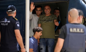 Three men stand inside a police van and one stands just in front of them. Two of the men are waving to crowds off-camera. Police officers stand facing the men with their backs to the camera.