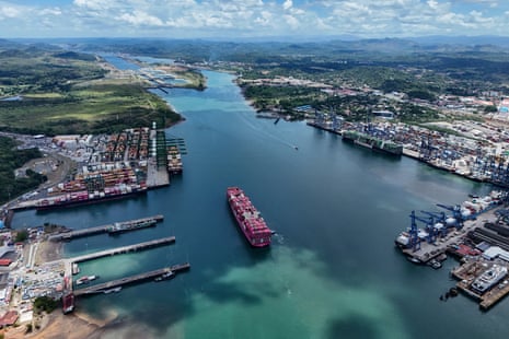 Aerial view of a red container ship entering the Panama Canal between busy port terminals
