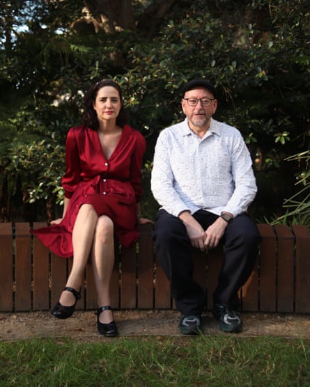 A woman with black wearing ruby-coloured satin dress and black mary-jane shoes sits on a bench outdoors next to a man wearing glasses and a pale-coloured long-sleeved shirt and black trousers