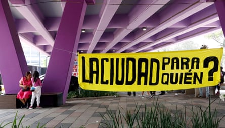 People stand near a large yellow banner reading ‘The city – for whom?’ in Spanish below an overpass painted purple