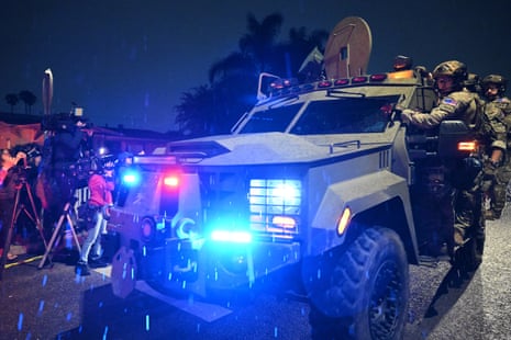 A large white tactical vehicle lights up the night with red and blue lights as uniformed FBI officers in full gear hang off the sides.