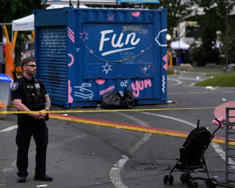 A police officer stands behind yellow tape near a blue shipping container decorated with 'Fun' and an abandoned stroller