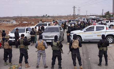 Armed men stand in a line across a road, with vehicles parked in front.
