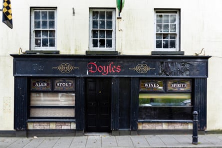 A closed-down pub with a black frontage and blinds down in its windows; its sign names it as Doyles in red lettering.