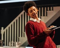 An actor smiles while looking at a device, headphones on. Leo Whitfield Harris in Ilford Boy by Danny at Donmar Warehouse, London.