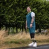 Full length shot of a 44-year-old man looking thoughtfully at the camera, leaning slightly to his left side away from the camera wearing running prostheses, white trainers and a white, green and yellow Team Australia top standing to side of a road on scorched grass with leafy trees behind him