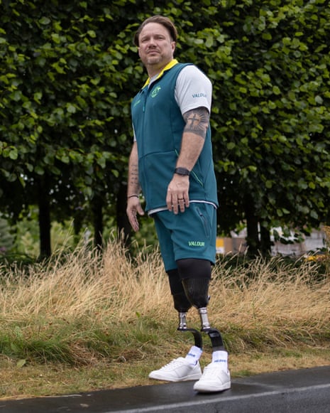 Full length shot of a 44-year-old man looking thoughtfully at the camera, leaning slightly to his left side away from the camera wearing running prostheses, white trainers and a white, green and yellow Team Australia top standing to side of a road on scorched grass with leafy trees behind him