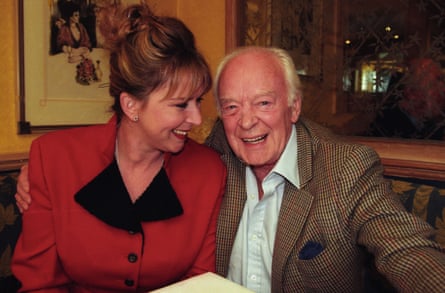 A women in a red jacket with black lapels sitting next to an old man with a tweed jacket, his arm round her