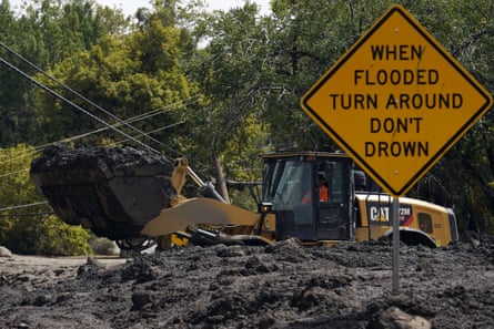 A bulldozer clears mud from a road. A sign in the foreground reads ‘When flooded turn around, don’t drown’.