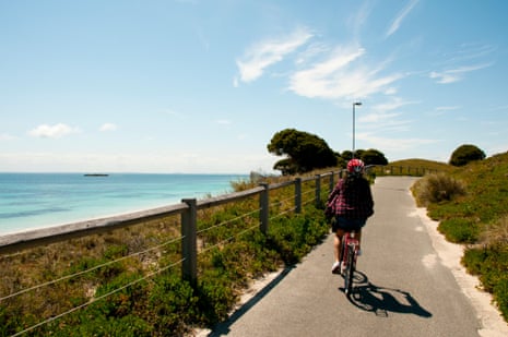 Back view of a cyclist riding on a paved footpath along a coastline, with a clear blue sky in the background