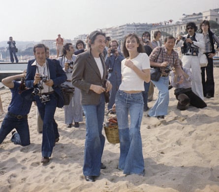 Jane Birkin and Serge Gainsbourg pose on the sand for photographers. They both wear flared denim jeans and white T-shirts, and she is carrying her basket