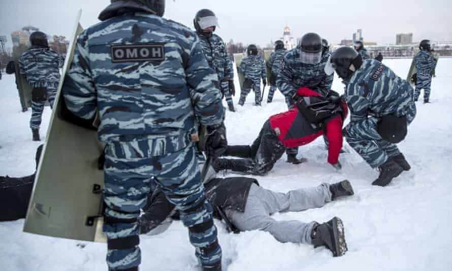 Police with riot shields and camouflage uniforms restrain two people lying down in the snow