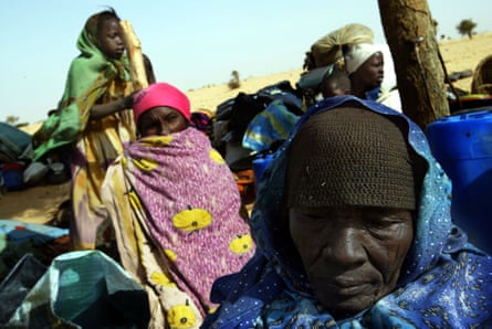 Sudanese women in traditional robes shelter under a tree in a desert landscape
