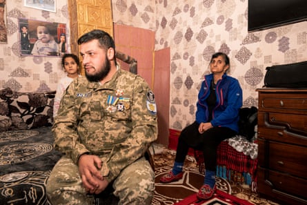 A man in army fatigues sits in a room with a baby picture on the wallpapered wall and rugs on the floor. A young girl is behind him and a woman sits to his left