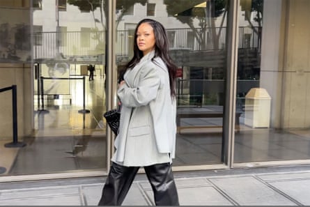 woman wearing grey coat and black pants looks at camera while walking in front of building