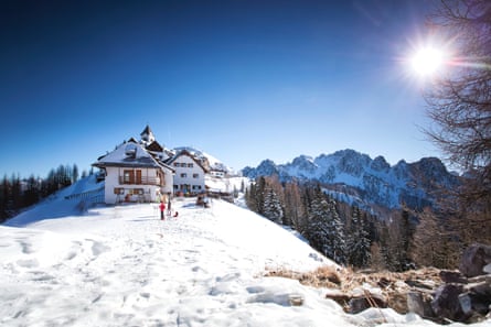 A couple of traditional chalets on top of a snowy hill with forest and mountain peaks behind